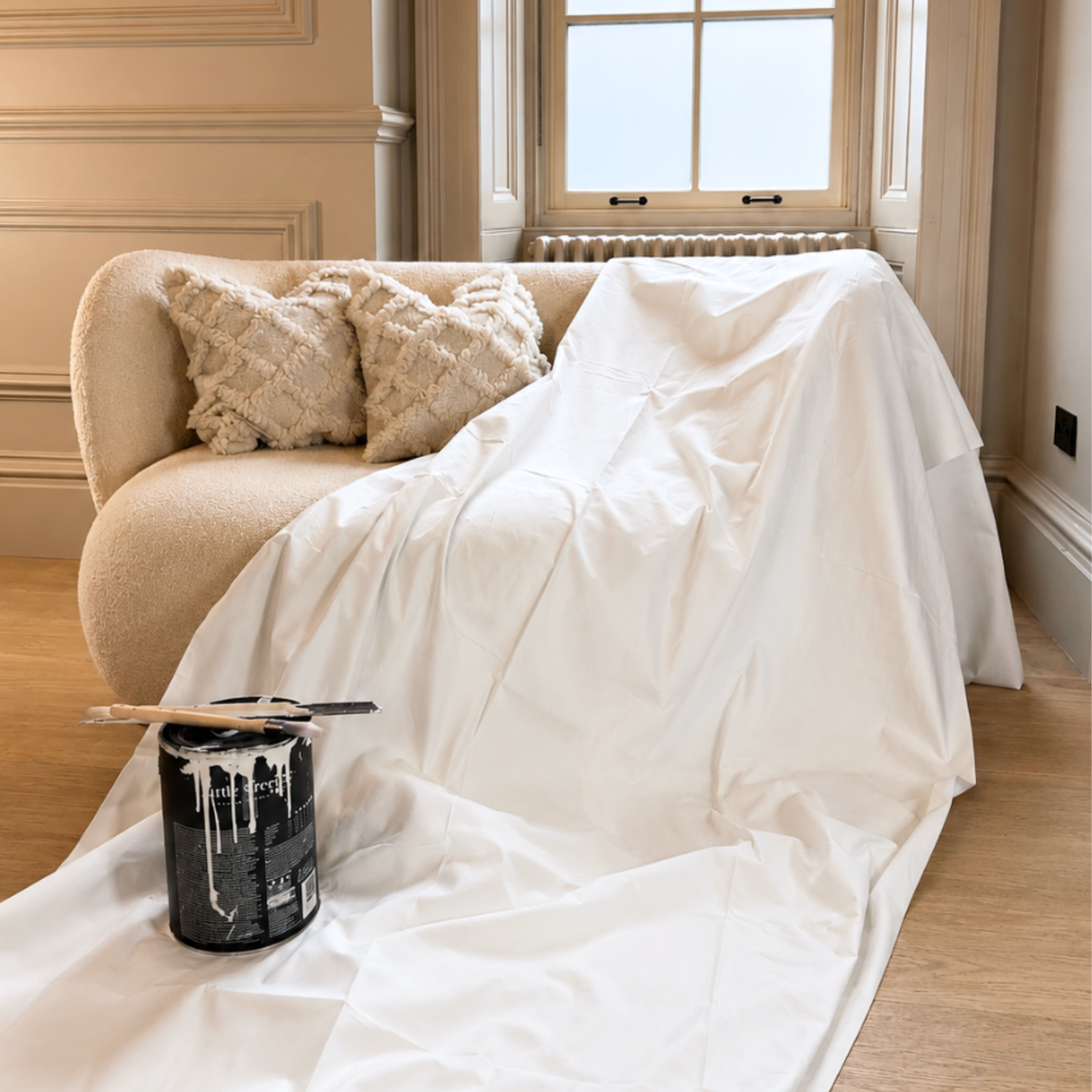 Beige sofa with white dust sheet and textured pillows in a room with a window.